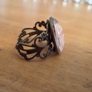 K&ouml;nnte beinhalten: Ein Ring mit einem filigranen, verzierten Band aus bronzefarbenem Metall und einem rosafarbenen Kamee in der Mitte.