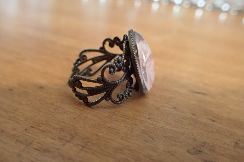 K&ouml;nnte beinhalten: Ein Ring mit einem filigranen, verzierten Band aus bronzefarbenem Metall und einem rosafarbenen Kamee in der Mitte.