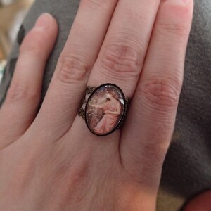 K&ouml;nnte beinhalten: Ein Ring mit einem ovalen Kamee, das eine Frau in einem rosa Kleid zeigt. Der Ring ist aus Bronze und hat ein filigranes Design.