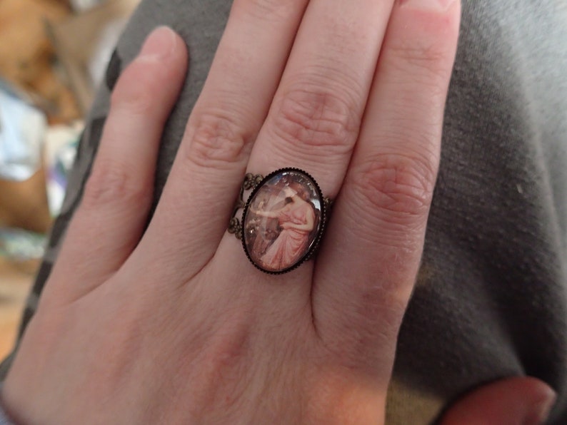 K&ouml;nnte beinhalten: Ein Ring mit einem ovalen Kamee, das eine Frau in einem rosa Kleid zeigt. Der Ring ist aus Bronze und hat ein filigranes Design.