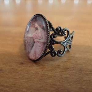 K&ouml;nnte beinhalten: Ein Bronzering mit einem ovalen Kamee, das eine Frau in einem rosa Kleid zeigt. Der Ring hat ein zartes, verziertes Design.