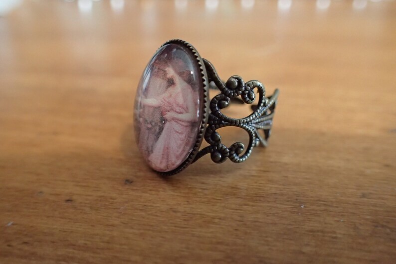 K&ouml;nnte beinhalten: Ein Bronzering mit einem ovalen Kamee, das eine Frau in einem rosa Kleid zeigt. Der Ring hat ein zartes, verziertes Design.