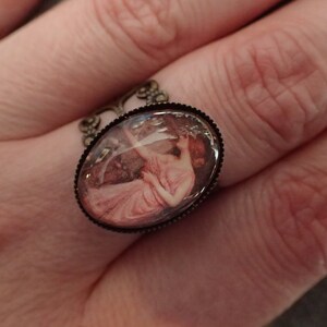 K&ouml;nnte beinhalten: Ein Ring mit einem Kamee, der eine Frau in einem rosa Kleid zeigt. Der Kamee ist in einem ovalen bronzefarbenen Rahmen mit einem dekorativen Filigranmuster gefasst.