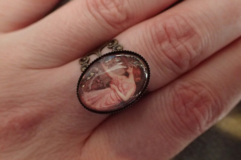 K&ouml;nnte beinhalten: Ein Ring mit einem Kamee, der eine Frau in einem rosa Kleid zeigt. Der Kamee ist in einem ovalen bronzefarbenen Rahmen mit einem dekorativen Filigranmuster gefasst.