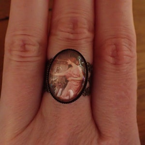 K&ouml;nnte beinhalten: Ein Ring mit einem Kamee, das eine Frau in einem rosa Kleid zeigt. Das Kamee ist in einem silberfarbenen Metallrahmen gefasst.