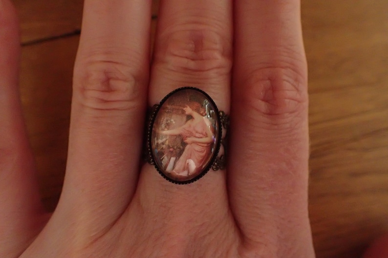 K&ouml;nnte beinhalten: Ein Ring mit einem Kamee, das eine Frau in einem rosa Kleid zeigt. Das Kamee ist in einem silberfarbenen Metallrahmen gefasst.