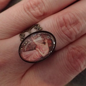 K&ouml;nnte beinhalten: Ein Ring mit einem ovalen Kamee, das eine Frau in einem rosa Kleid zeigt. Der Ring ist aus Bronze und hat ein filigranes Design.