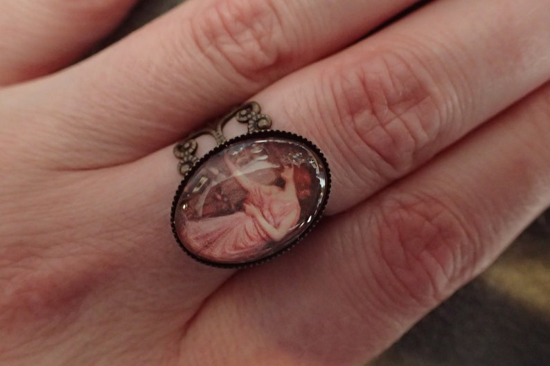K&ouml;nnte beinhalten: Ein Ring mit einem ovalen Kamee, das eine Frau in einem rosa Kleid zeigt. Der Ring ist aus Bronze und hat ein filigranes Design.