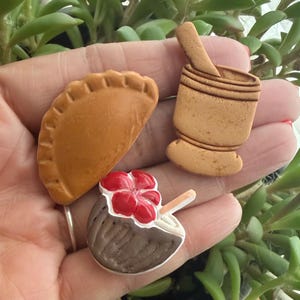 May include: Three small decorative items including a tan crescent-shaped pastry with crimped edges, a tan mortar and pestle set, and a dark brown coconut shell half topped with a bright red flower and a small light-colored straw or stick.
