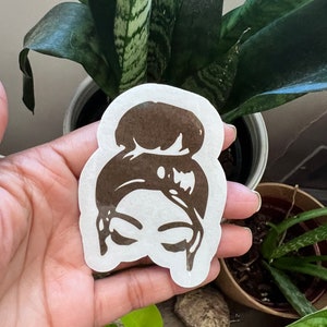 May include: A brown silhouette sticker of a person with a bun hairstyle. The sticker is on a white background and is being held by a hand.