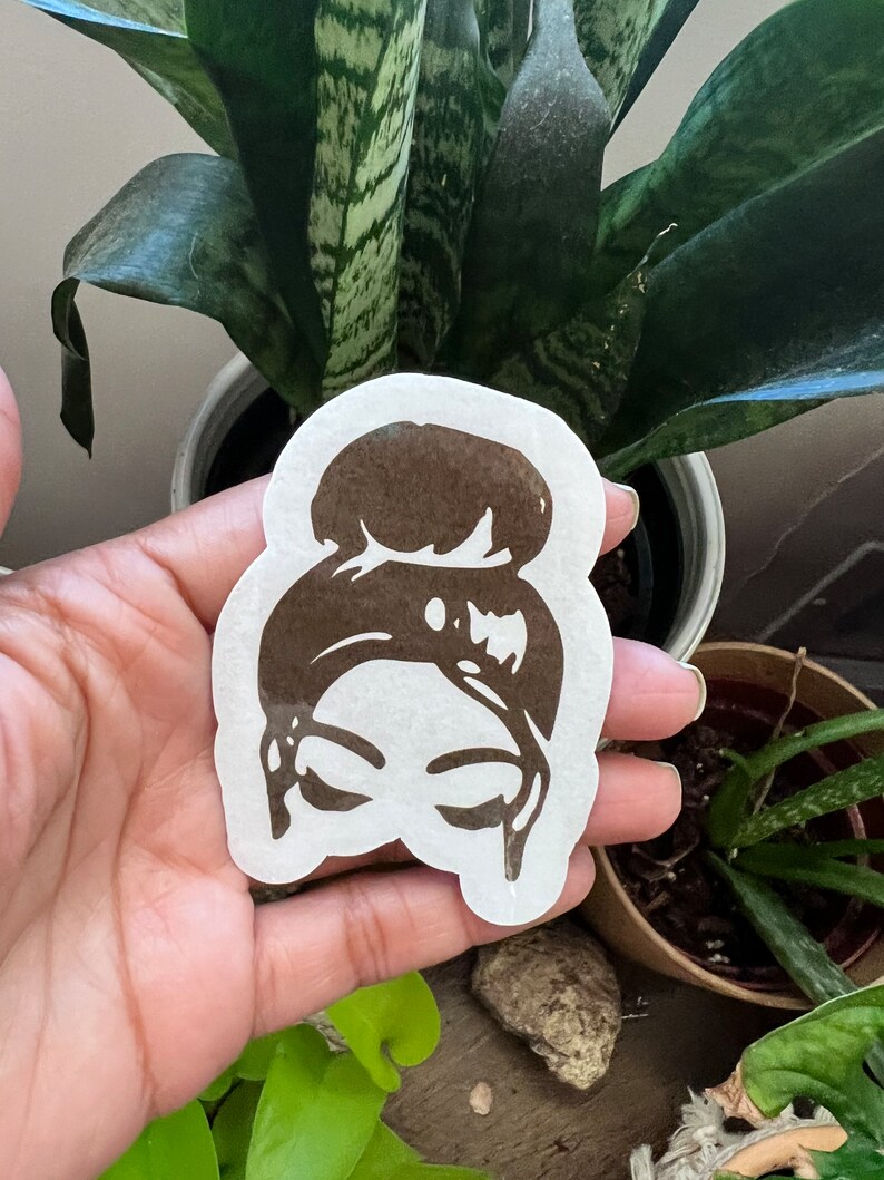 May include: A brown silhouette sticker of a person with a bun hairstyle. The sticker is on a white background and is being held by a hand.