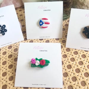 May include: Four white cards with the text "Melissa Creates" printed on them. Each card features a different handmade clay pin: a black and white abstract design, a red, white, and blue Puerto Rican flag, a black and white abstract design, and a cluster of pink, red, and white flowers.