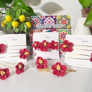 Op de afbeelding: Rode bloemen haaraccessoires, waaronder haarspeldjes en haarclips, met gele centra. De accessoires worden gepresenteerd op witte kaarten met de tekst "Melissa Creates". Een decoratief "Puerto Rico"-bord staat op de achtergrond.