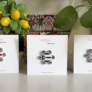 May include: Three enamel pins on white cards. The pins have a flower-like design with the flag of Puerto Rico. The cards have the text "Melissa Creates" and "Handmade in New York". The background includes plants and a decorative tile.