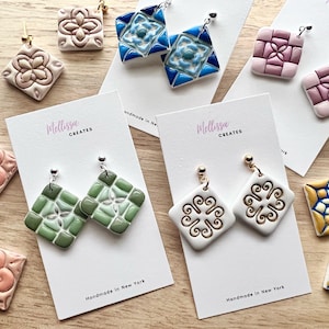May include: Assortment of handmade ceramic tile earrings in various colors and patterns. The earrings are displayed on white cards with the text "Mellissa Creates" and "Handmade in New York".