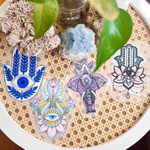 May include: Four hamsa hand stickers with different designs. The stickers are on a white surface with a woven pattern. The hamsa hands are blue, pink, yellow, and purple with an evil eye in the center of each hand.