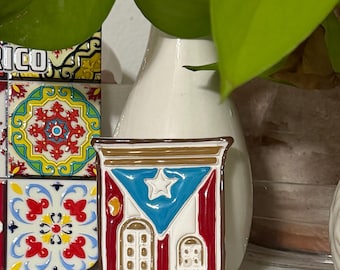 May include: A rectangular, colorful magnet featuring the Puerto Rican flag design. The magnet has a blue triangle with a white star, red stripes, and a white background with architectural details. The magnet is framed in brown.