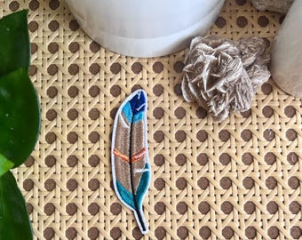 Könnte beinhalten: Ein bunter gestickter Federn-Patch mit blauen, orangefarbenen und braunen Akzenten. Der Patch befindet sich auf einer geflochtenen Weidenoberfläche.