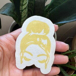 May include: A gold-colored sticker with a silhouette of a woman's head with a bun hairstyle.