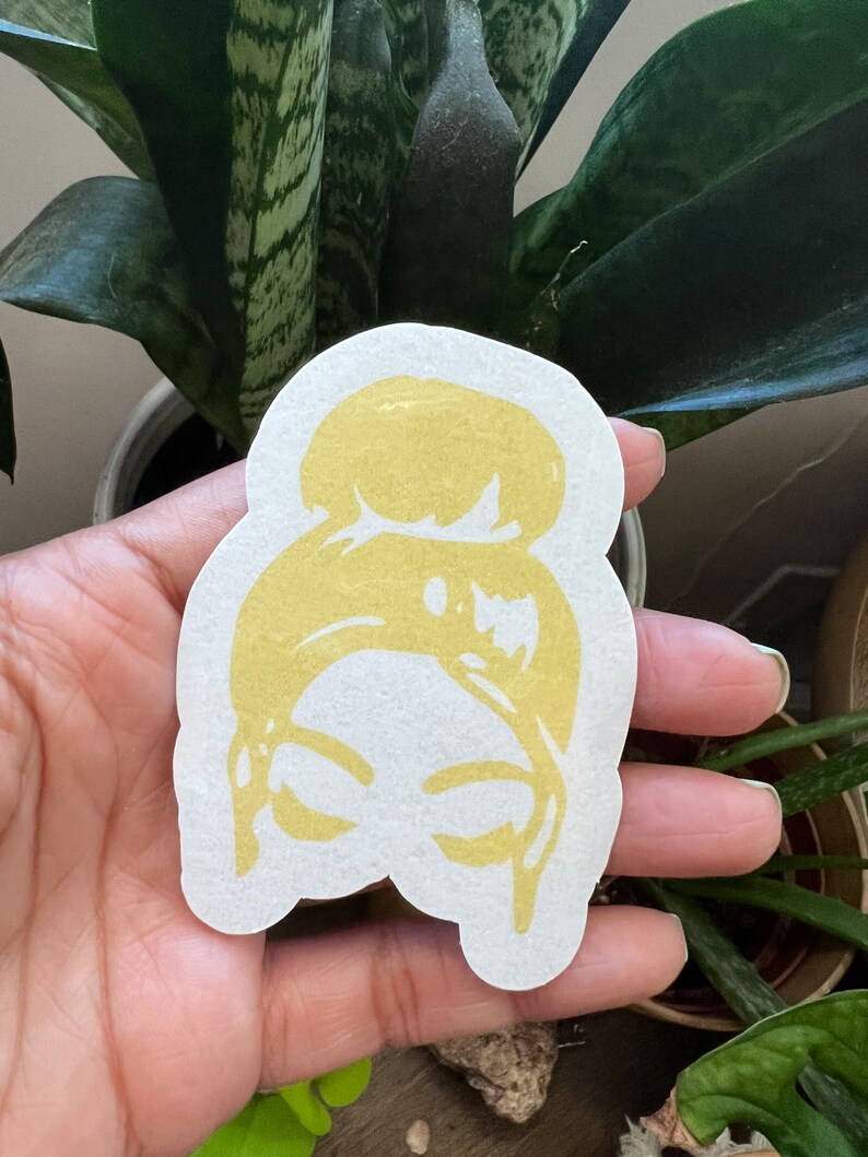 May include: A gold-colored sticker with a silhouette of a woman's head with a bun hairstyle.