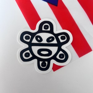 May include: A black and white sticker of the  "Sol de Borinquen" symbol, a stylized sun with a face, representing Puerto Rico.