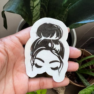 May include: Black and white sticker of a woman with her hair in a bun. The woman's eyes are closed and she is looking down.