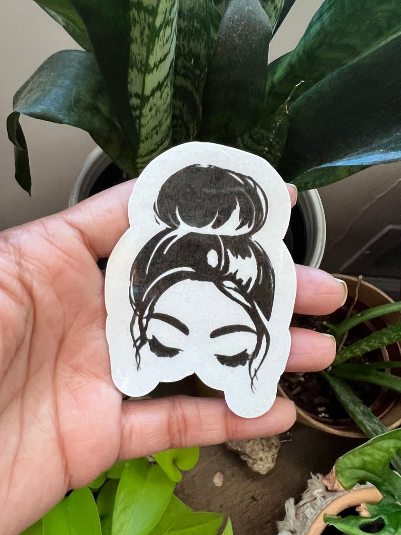 May include: Black and white sticker of a woman with her hair in a bun. The woman's eyes are closed and she is looking down.