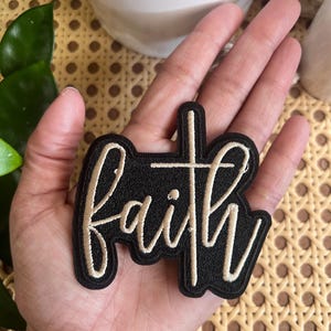 Puede incluir: Un parche de tela negro con la palabra "faith" bordada en hilo beige. La palabra está estilizada con una cruz en el centro de la "a".
