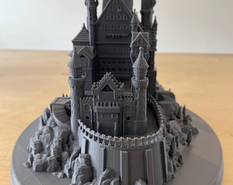May include: A 3D printed model of a dark gray castle with a tall tower and a surrounding wall. The castle is set on a rocky base and is surrounded by a circular base.