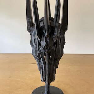 May include: A black 3D printed crown with a pointed, jagged design. The crown is mounted on a black circular base.