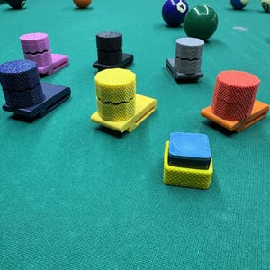 May include: A collection of multi-colored pool cue chalk holders with textured cylindrical tops and clip bases, arranged on a green felt billiard table alongside various numbered billiard balls scattered across the surface of the table.