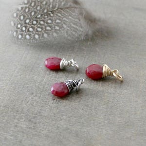 May include: Three small, faceted, teardrop-shaped red gemstones are wire-wrapped in silver and gold. The gemstones are arranged on a gray surface, with a large, speckled feather in the background. The gemstones are likely jewelry components.