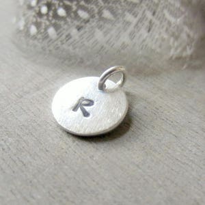 May include: A small, round silver pendant with the letter "R" engraved on its surface. The pendant has a textured finish and a small loop at the top for attaching to a chain or bracelet. The background is a soft, blurred gray.