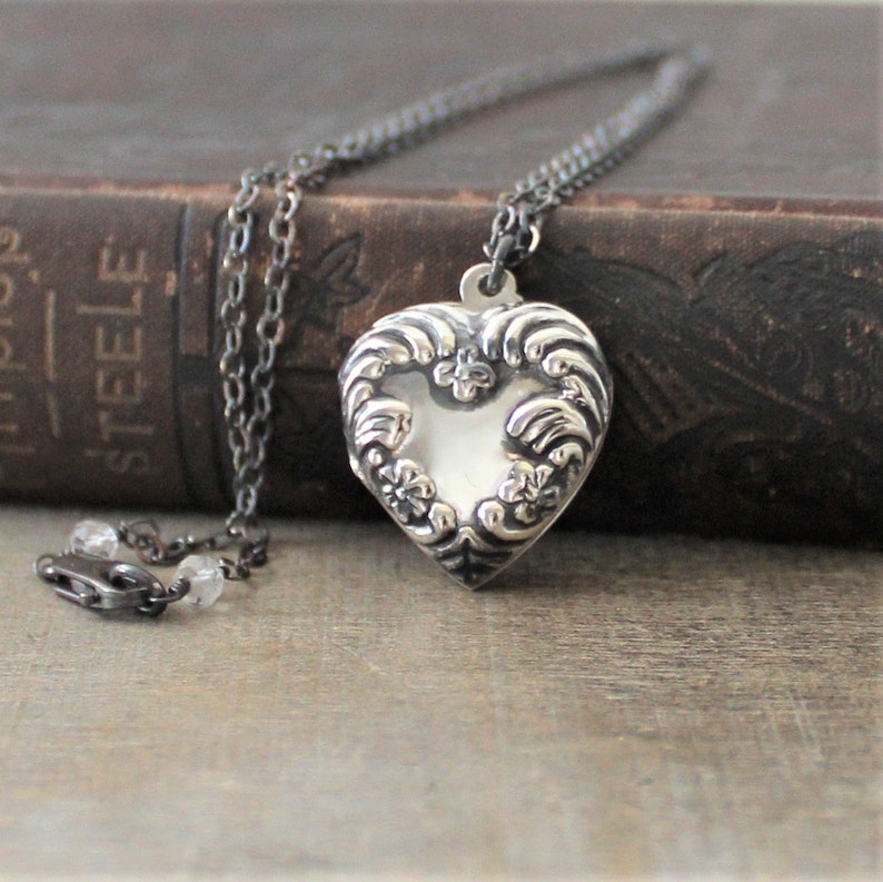 May include: A silver heart-shaped locket necklace with a detailed floral design. The locket is on a black chain and is resting on a brown book.