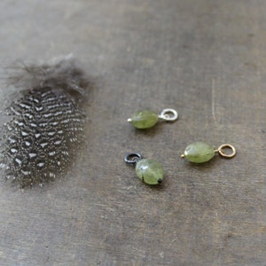 May include: Three green gemstone pendants with silver and gold accents. The pendants are on a wooden surface.