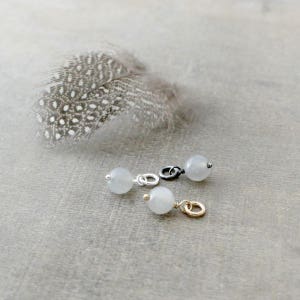 May include: Three silver, gold, and black charms with a white moonstone bead on each. The charms are on a light gray surface.
