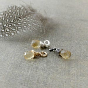 May include: Three gold and silver wire wrapped gemstone charms with a teardrop shaped citrine stone. The charms are on a light brown surface.