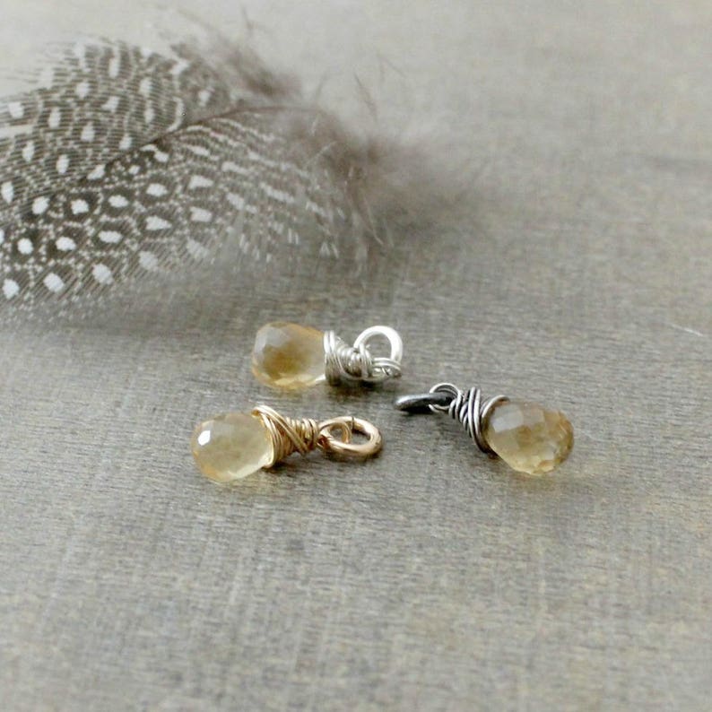 May include: Three gold and silver wire wrapped gemstone charms with a teardrop shaped citrine stone. The charms are on a light brown surface.