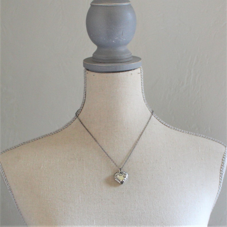 May include: A silver heart-shaped locket pendant on a silver chain necklace. The locket is on a white mannequin bust.