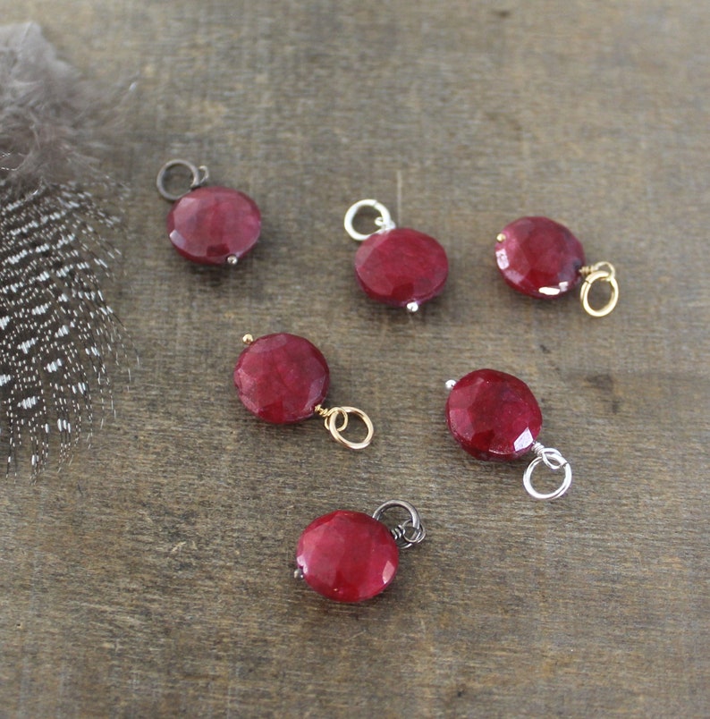 May include: Six red gemstone pendants with silver and gold jump rings. The gemstones are round and faceted. The pendants are on a wooden surface.