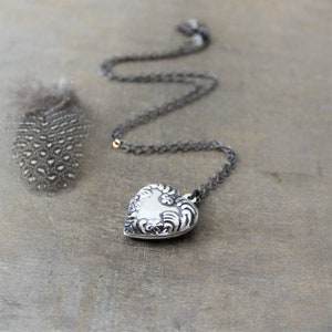 May include: A silver heart-shaped locket necklace with a detailed floral design. The chain is black and the locket is hanging on a wooden surface.