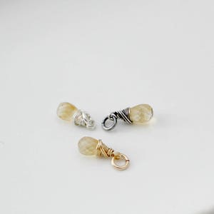May include: Three small teardrop-shaped gemstone charms with wire wrapping. The charms are made with a light yellow gemstone and silver or gold wire.