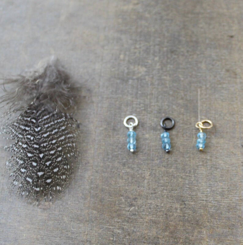 May include: Three charm pendants with blue gemstone beads on silver, black, and gold chains. The charms are on a wooden surface.