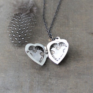 May include: A silver heart-shaped locket necklace with an intricate floral design. The locket is open, revealing the inside. The necklace is on a black chain.