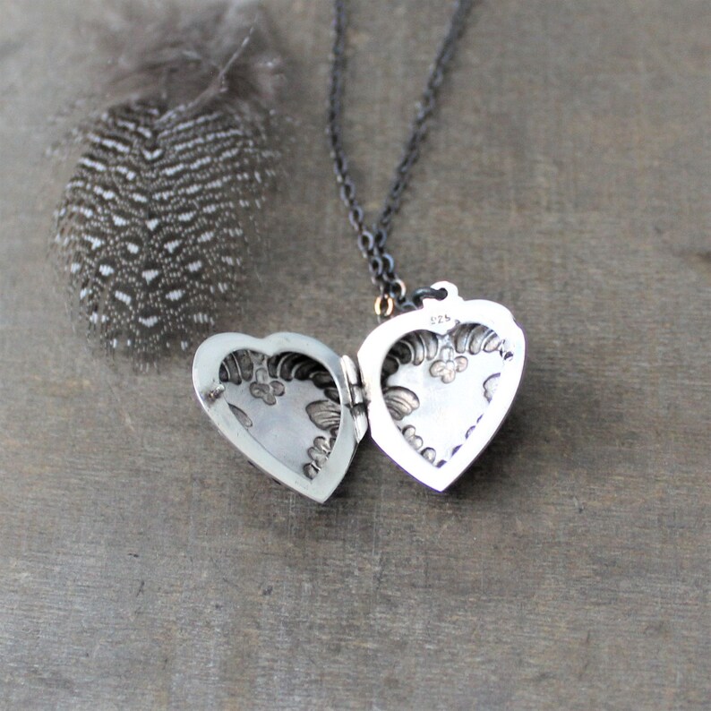 May include: A silver heart-shaped locket necklace with an intricate floral design. The locket is open, revealing the inside. The necklace is on a black chain.