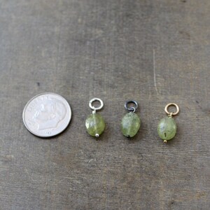 May include: Three green gemstone pendants with different colored metal loops. The pendants are oval shaped and have a faceted finish. The loops are silver, black, and gold.