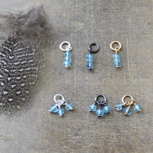 May include: Six different charms featuring blue topaz gemstones. Three charms have three gemstones each, and three charms have one gemstone each. The charms are available in silver, gold, and black finishes.