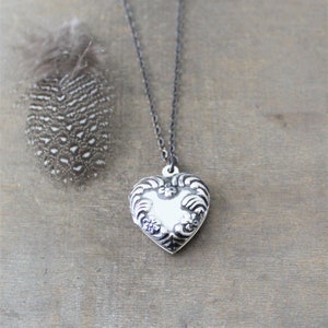 May include: A silver heart-shaped locket pendant on a black chain necklace. The locket has an ornate design with floral details.
