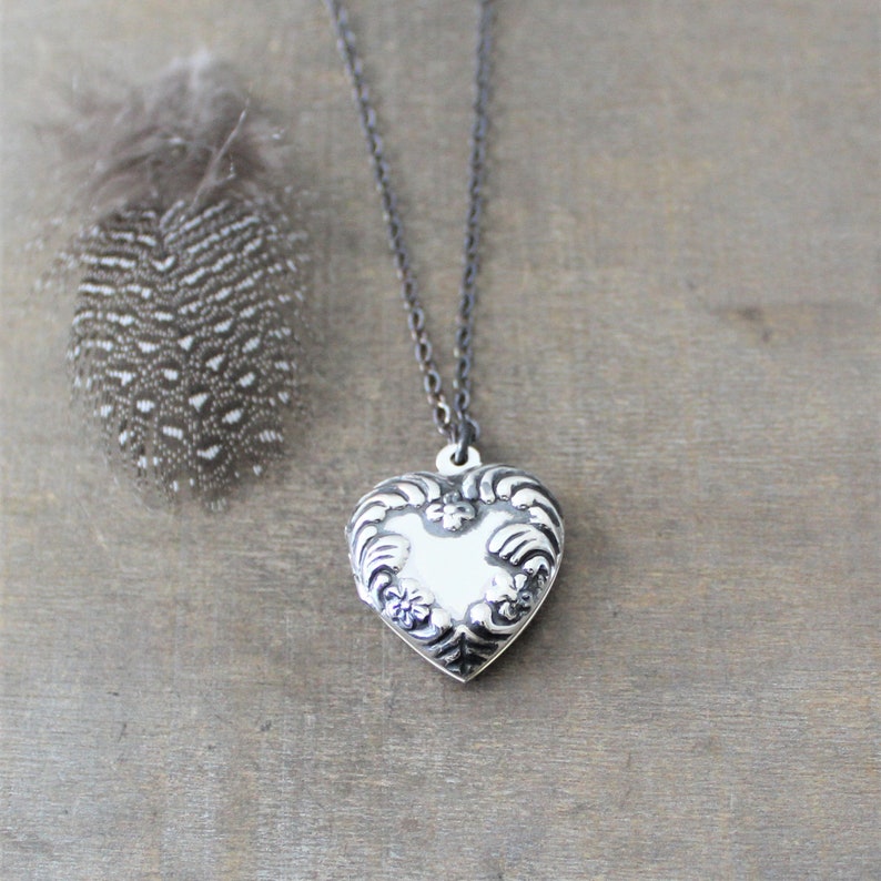 May include: A silver heart-shaped locket pendant on a black chain necklace. The locket has an ornate design with floral details.