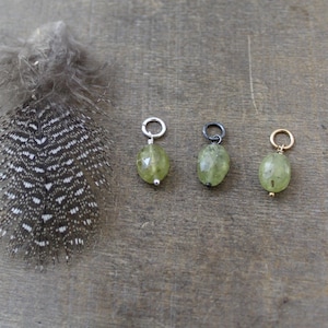 May include: Three oval-shaped green gemstone charms with different colored metal jump rings. The charms are on a wooden surface.